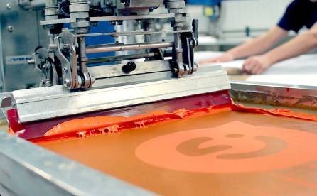 A machine is screen printing orange inks on substrates.
